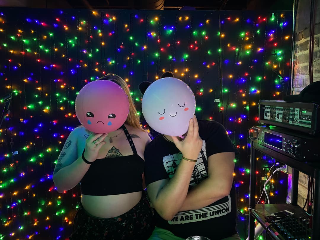 Two DJs holding up happy and sad face balloons.